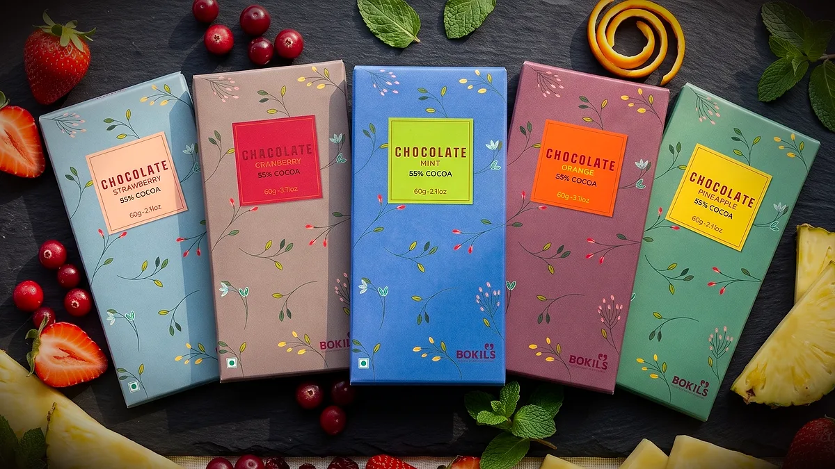 Five Bokils chocolate bars in pastel wrappers — Strawberry, Cranberry, Mint, Orange and Pineapple — with fruit on slate
